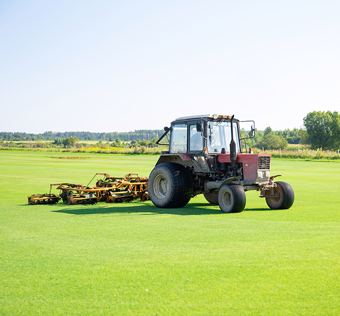 SERVICES – GREENBELT Tractor with a nozzle for care of a field lawn for a horse polo in work. The cut-off grass takes off from mechanical parts of a nozzle. Green flat field. Sky with beautiful clouds on background